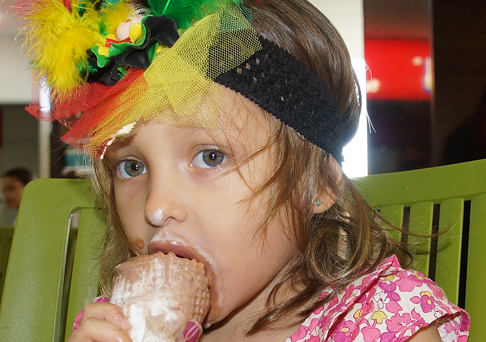 Niña colombo americana con una banda de carnaval en su cabeza comiendo un cono de helado